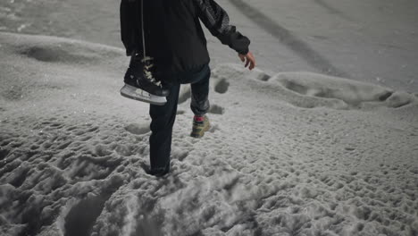 back view of skater stepping carefully down snowy stairs holding black ice skate boot, surrounded by thick layers of snow and rough winter textures under soft lighting on cold night
