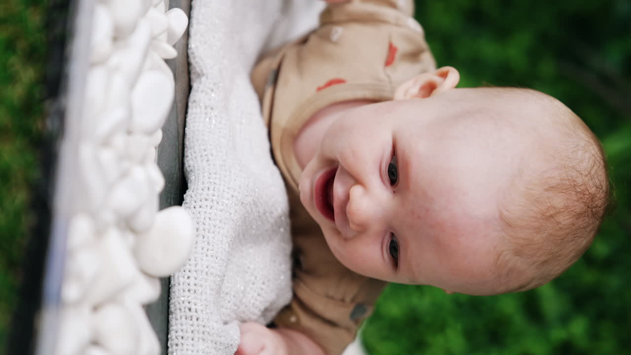 Cute Caucasian infant lying on his belly and holding head up. Baby boy smiling adorably. Close up. Vertical screen. Greenery in blur at backdrop.