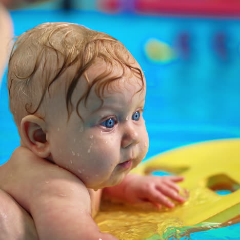 beautiful blue-eyed baby boy swims on the board in the swimming pool. female coach supports the kid.