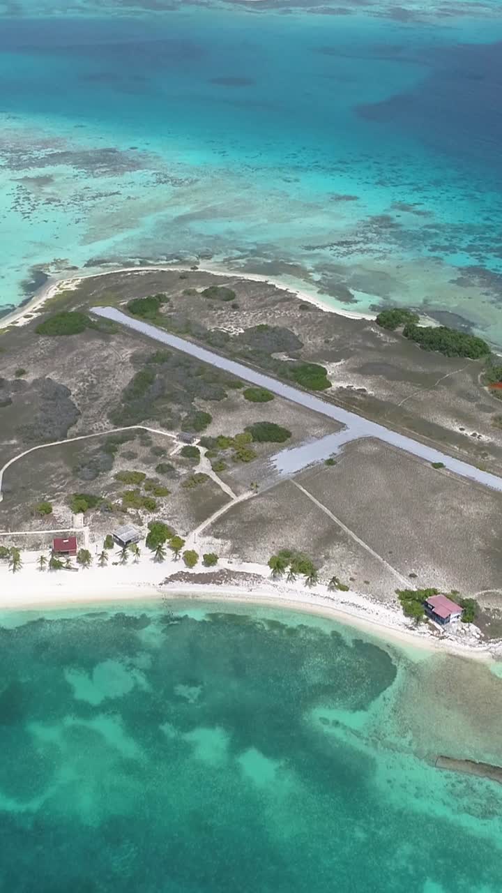 Drone shot remote tropical island surrounded by vivid turquoise and deep blue waters. Small landing strip cutting across dry coastal terrain, with scattered vegetation and beachfront structures