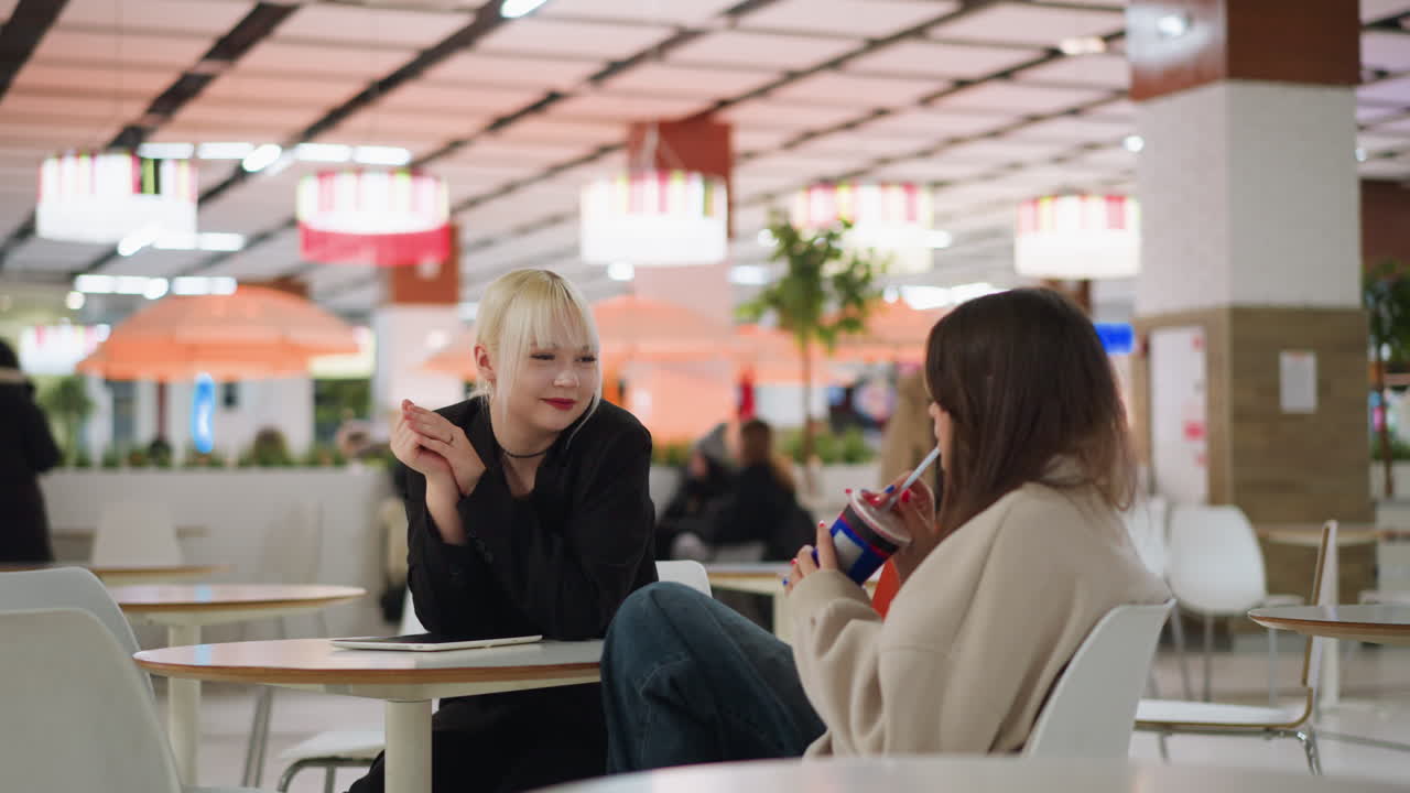 Two young women sit together at table indoors as one holds drink cup with straw, sharing beverage while smiling and enjoying relaxed conversation, with bright lights and blurred background