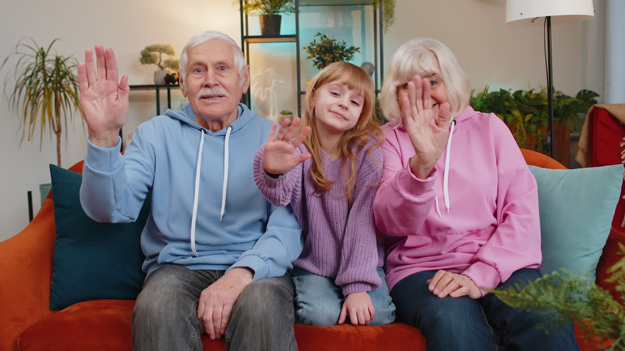 Child girl granddaughter with grandparents bloggers waves hand hello hi relaxing making on home sofa
