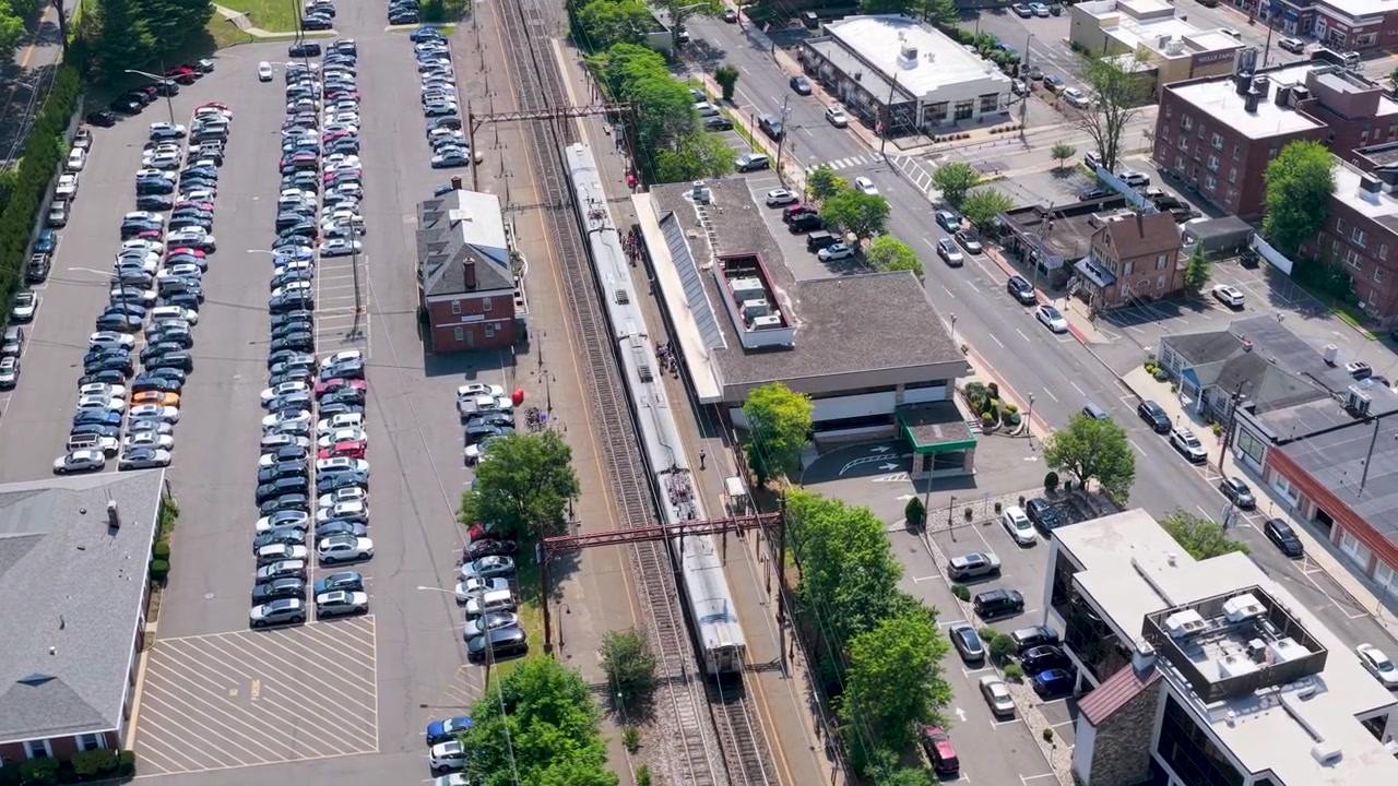 Bovenaanzicht van een forensentrein die aankomt op station Millburn in New Jersey, met het drukke perron, de aangrenzende volle parkeerplaats en het omliggende voorstedelijke centrumgebied met verkeer