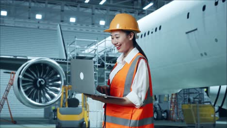 vista lateral de una ingeniera asiática con casco de seguridad trabajando en una computadora portátil mientras está de pie con aviones en el hangar