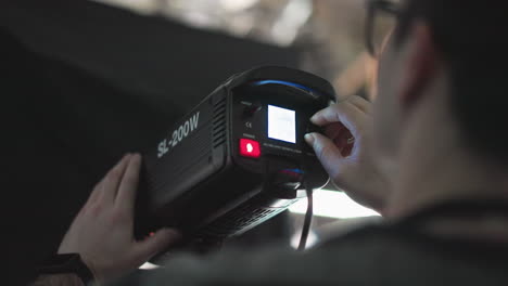 technician adjusting power switch and brightness knob on sl-200w led studio light mounted on bracket, cables visible, set against blue lit backdrop during broadcast production setup