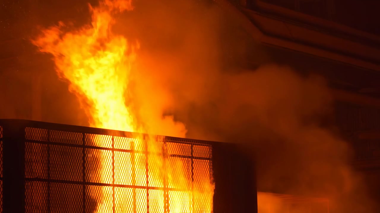 Tall flames rise, twist, and briefly recede before flaring again behind a rigid metal grid, while heavy smoke drifts across the dim ceiling and orange light fills the scene.