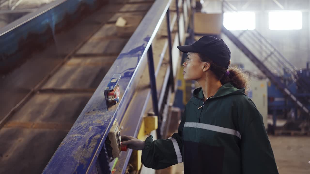 Young african american woman checking conveyor belt with push buttons at waste recycling plant. Pollution control