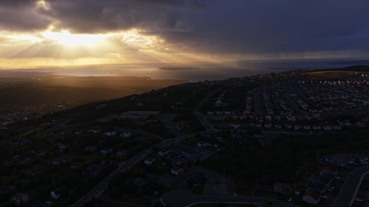 Cinematic aerial footage capturing god rays piercing clouds above Newfoundland's Conception Bay. Sunbeams light rugged coastline and shimmering water below, revealing a serene seascape
