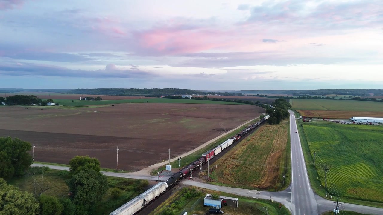 A drone captures a long freight train rolling past a quiet countryside road crossing near a grain facility as vivid pink sunset clouds stretch across the sky above rolling farmland.