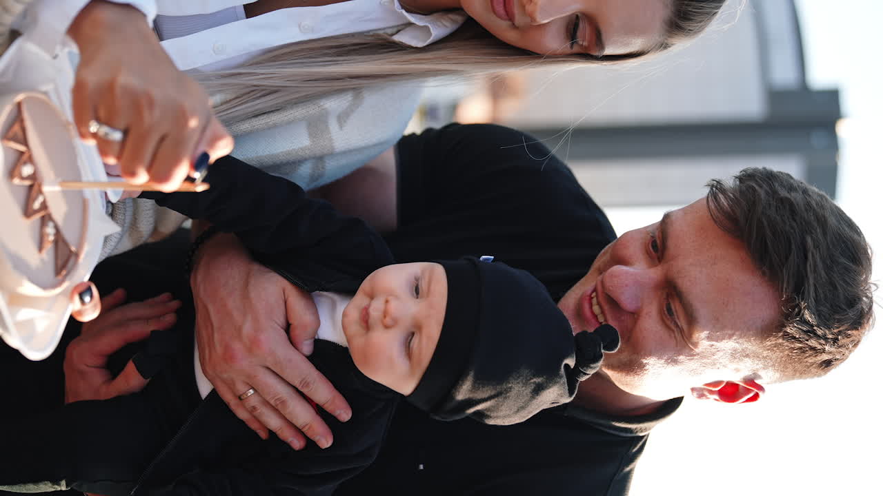 Caucasian smiling man with a baby in hands. Long-haired woman tries to light a candle on the cake. Vertical screen.