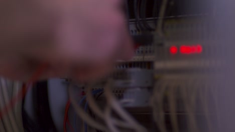 a focused technician, with a blurred face, diligently working on an active server rack, bathed in the glow of three red lights, in a room filled with machinery and equipment, caught by a static camera