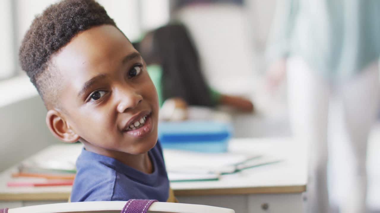Video of happy african american boy sitting at desk during lesson in classroom