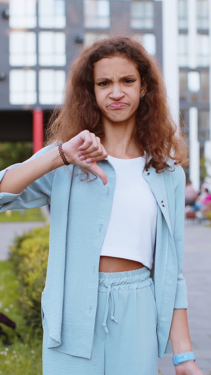 Upset preteen child girl showing thumbs down sign disapproval dissatisfied bad work mistake in city