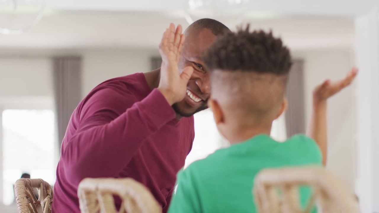 Happy african american man and his son sitting at table and high-fiving