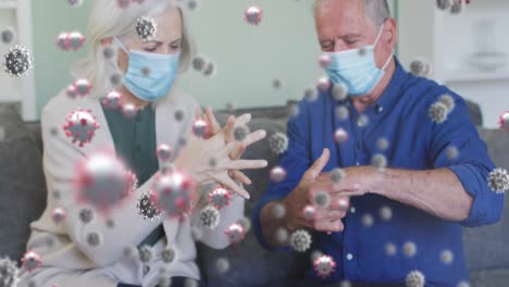 animation of virus cells over senior caucasian couple disinfecting hands at home with face masks