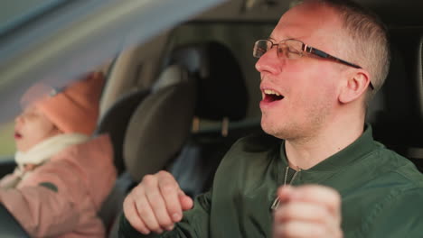 a man is seated in the driver's seat of a car, wearing a green jacket and glasses, smiling and gently dancing to music while singing. beside him, a young girl, dressed in a pink beanie and jacket