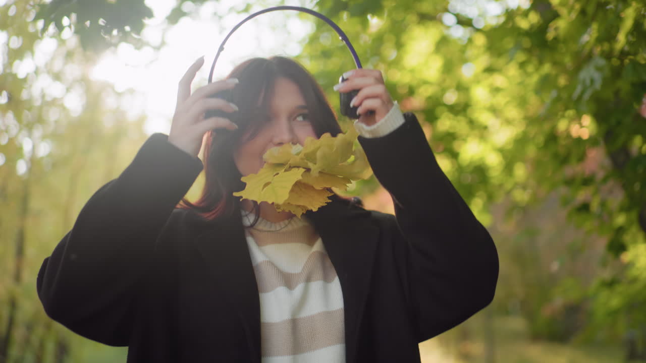 Autumn explorer standing with bunch of yellow leaves in mouth while wearing headphones, smiling and moving body with excitement under warm sunlight surrounded by forest trees and seasonal foliage