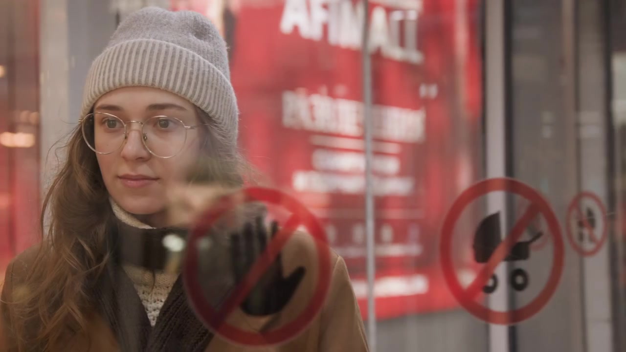 Mujer blanca pasando puertas de cristal, con reflejos de la estación e iconos de seguridad en el vidrio. Viste gorro de punto y abrigo de invierno, con luces de fondo borrosas. Muestra una expresión introspectiva y gestos cercanos, en la entrada de un transporte público.