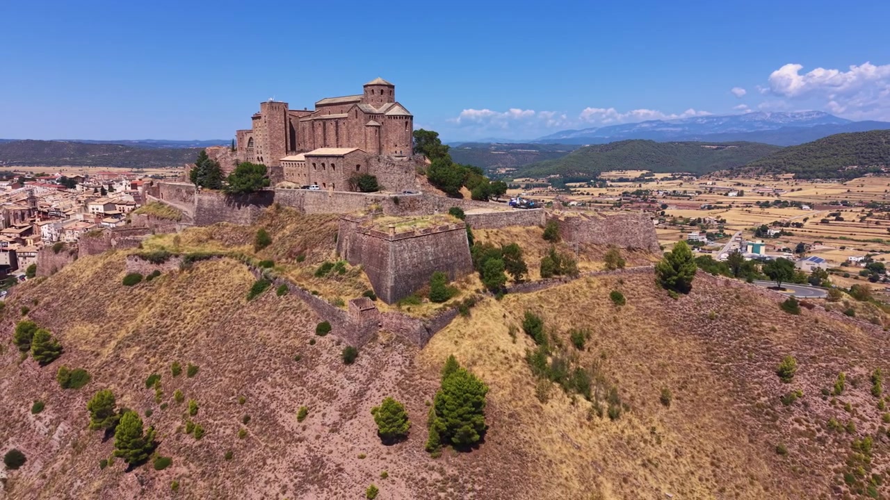 Castello di Cardona arroccato su una collina rocciosa in Catalogna, Spagna, con mura medievali in pietra, la città circostante, valli verdi e catena montuosa sotto un cielo blu.