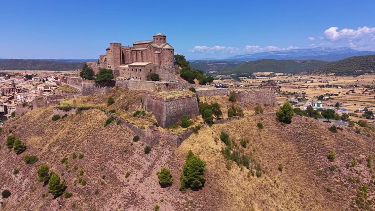 Castello di Cardona arroccato su una collina rocciosa in Catalogna, Spagna, con mura medievali in pietra, la città circostante, valli verdi e catena montuosa sotto un cielo blu.