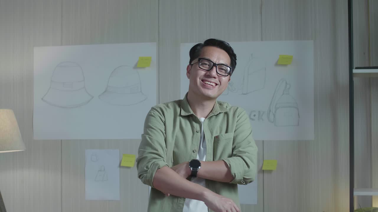 아시아 남자 디자이너가 팔을 고 벽에 있는 가방 디자인 스케치 종이 앞에 서서 카메라를 향해 미소 짓는다.