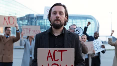 retrato de un hombre caucásico con barba sosteniendo un cartel de actuar ahora en una protesta con colegas de negocios multiétnicos en la calle