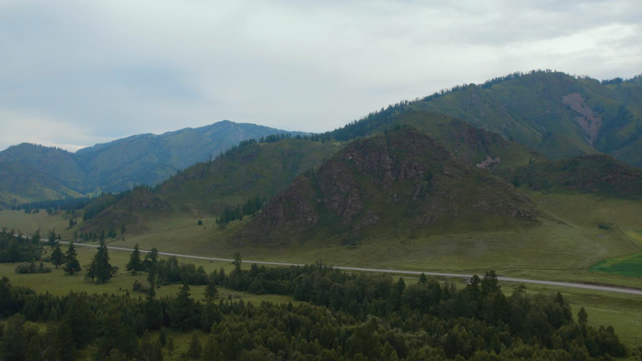 Riprese aeree con drone di un'ampia valle montana verde con colline boscose e una strada rurale sotto un cielo coperto nella Repubblica dell'Altai, Russia, un sereno paesaggio estivo.