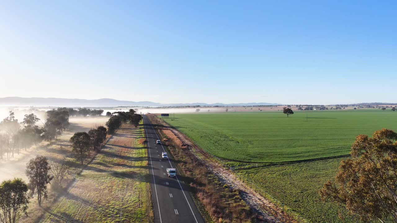 Aerial footage shows a car traveling along a country road bordered by green fields and trees, with morning fog and soft sunlight creating a tranquil atmosphere