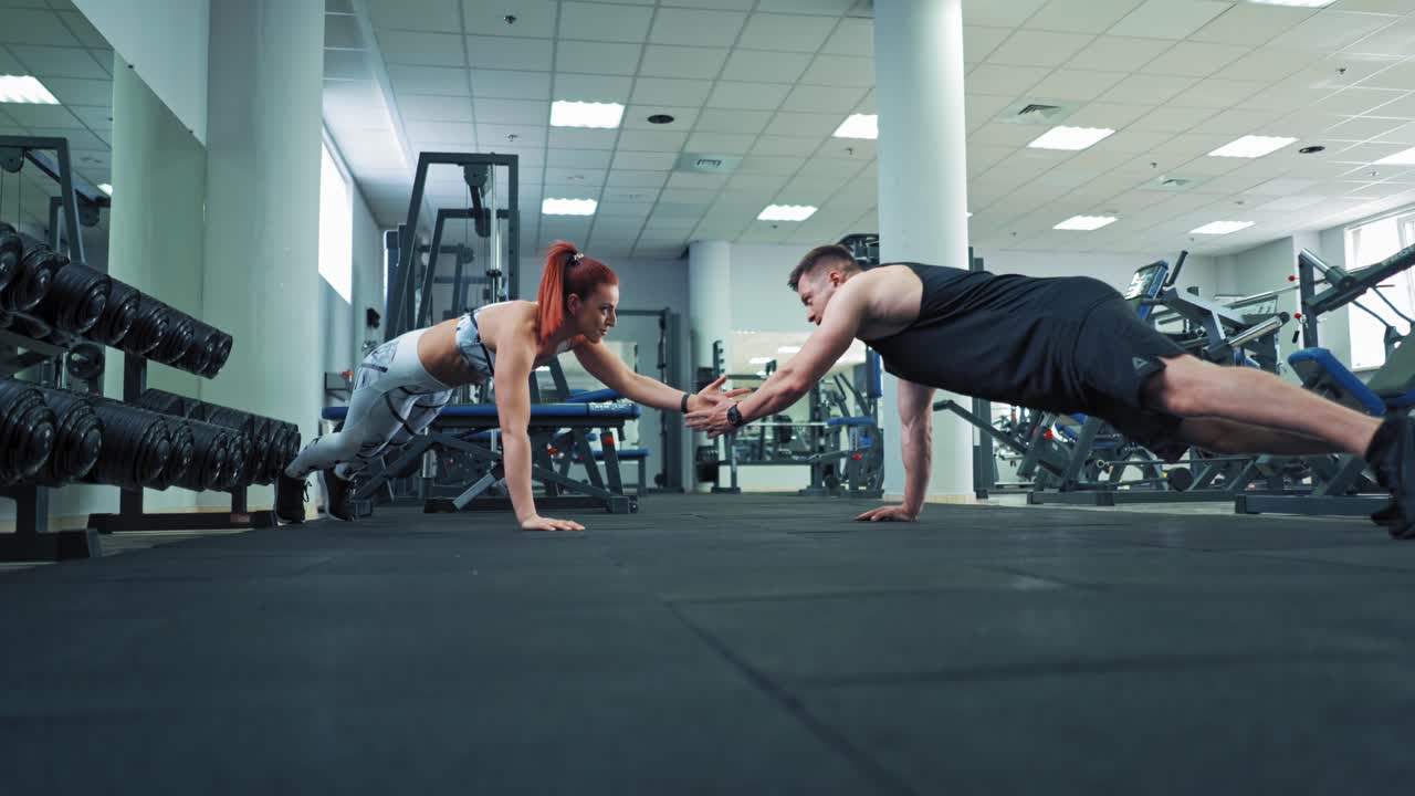 Muscular athletic man and beautiful fitness woman doing push ups exercise indoors. Healthy couple exercising on the floor one opposite other and giving five together in fitness center.