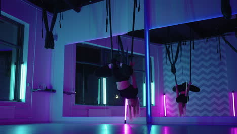 a woman does sports yoga in the air on a hammock in neon light performing graceful exercises and movements from stretching and dancing in slow motion.