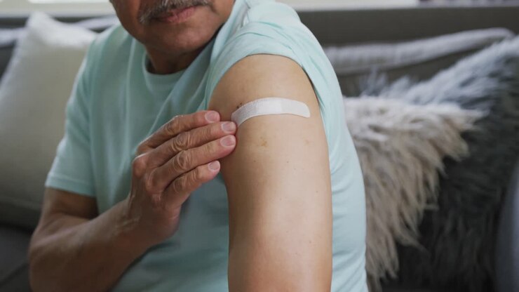Vaccinated elderly man touching bandage on arm while sitting by sofa at home
