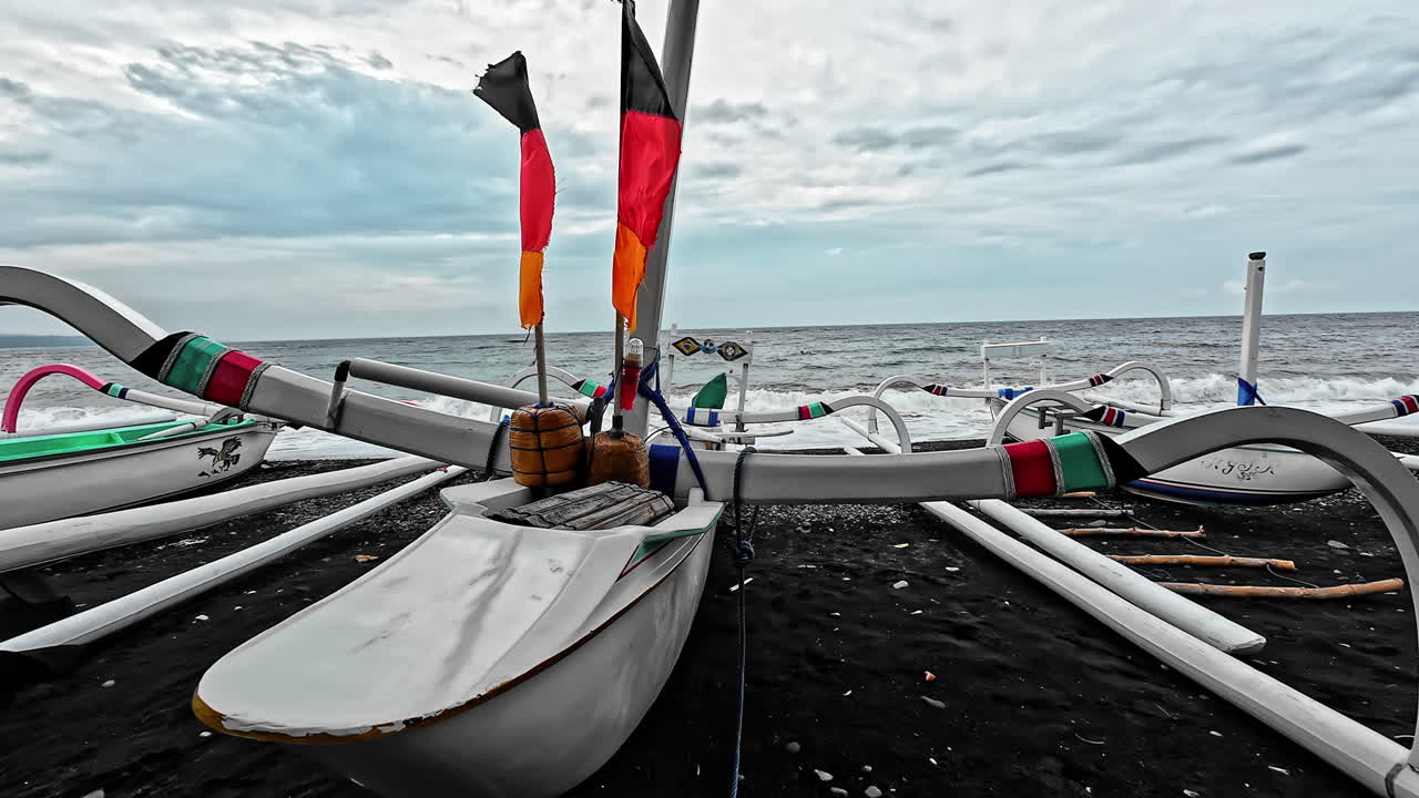Colorful Balinese jukung boats rest on a black sand beach facing the sea. Bali, Indonesia