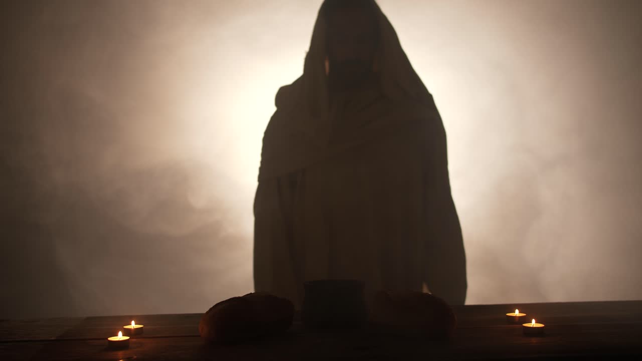 The silhouette of Jesus Christ standing near a table with Christian sacraments of bread and wine for last supper, communion, Easter symbolism. In cinematic slow motion with ambient light.