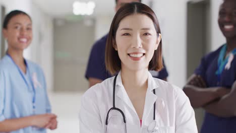 Video-portrait-of-smiling-asian-female-doctor-with-cancer-ribbon-in-hospital-corridor,-copy-space