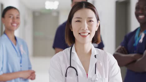 video portrait of smiling asian female doctor with cancer ribbon in hospital corridor, copy space