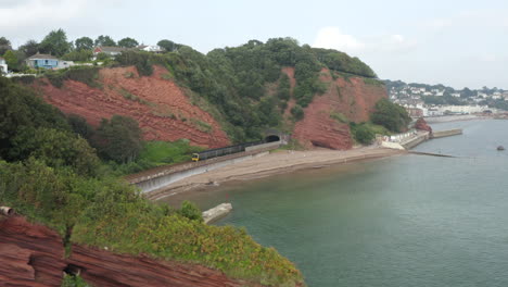 an aerial shot flying over red cliffs at dawlish with a train coming out of a tunnel on the devon coast with the beach and sea in shot