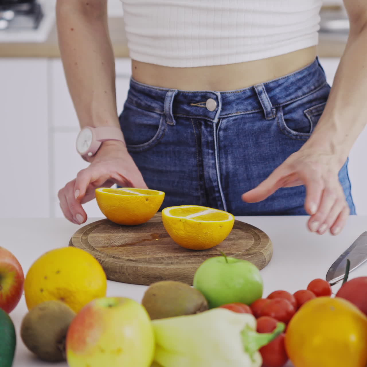 Woman with sharp knife cutting orange into two halves. Table full of fresh fruit and vegetables in the kitchen. Woman preparing healthy food for losing weight.