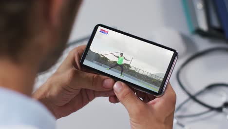 compuesto de un médico masculino viendo un evento de jabalina de atletismo en un teléfono inteligente