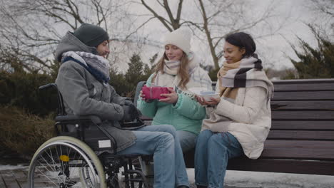 two women with small birthday cake and a gift giving a surprise to their disabled friend in wheelchair at urban park in winter