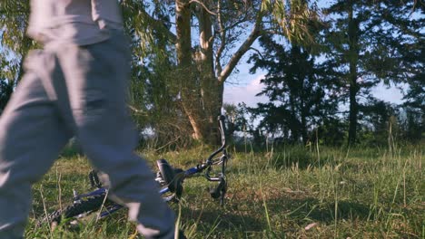 Caucasian-5-years-old-boy-trying-to-pick-up-his-fallen-bicycle-after-a-crash-in-the-fields,-forest-trees-in-the-background,-4k-shot