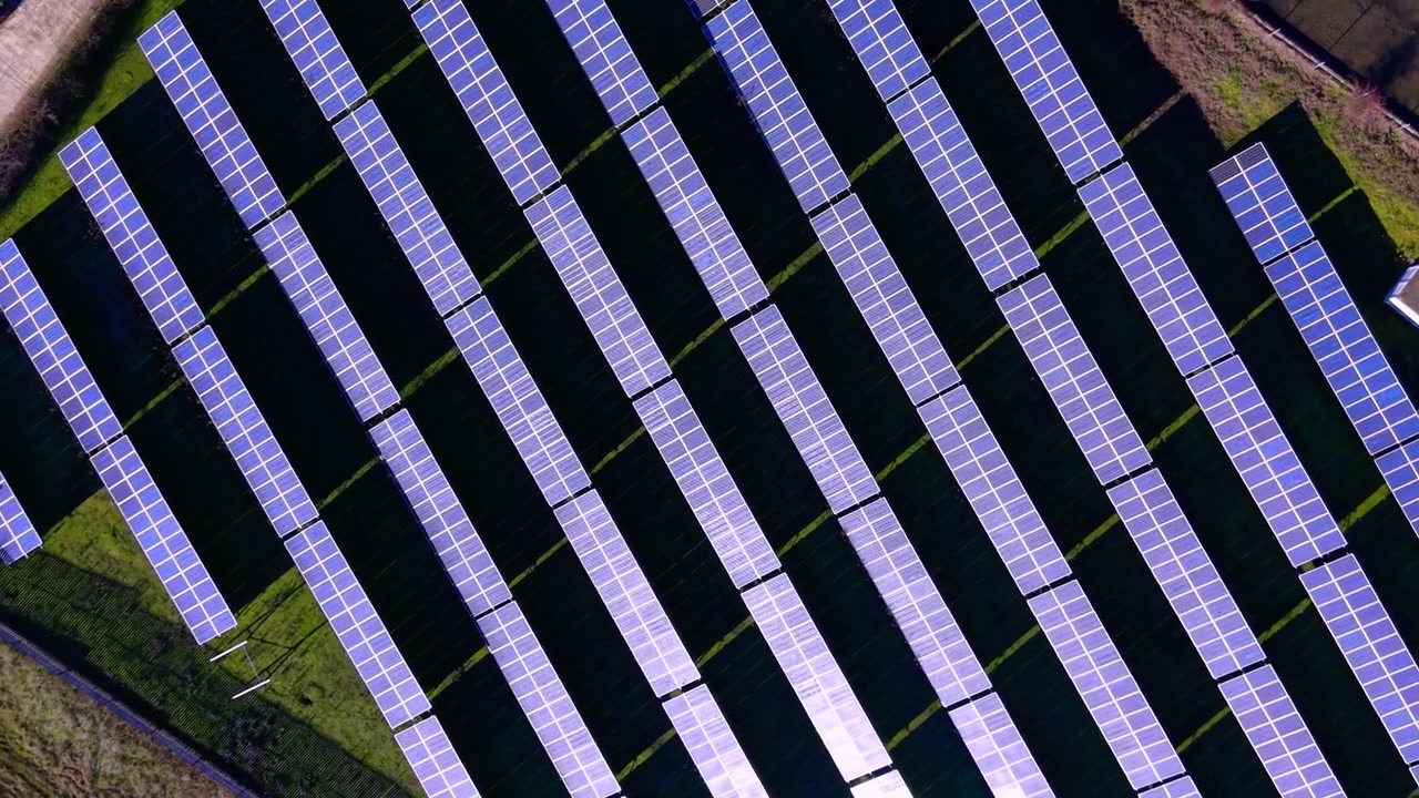 Vista dall'alto di un impianto di pannelli solari in file diagonali. File di moduli fotovoltaici riflettono luce viola e blu in un motivo a griglia.