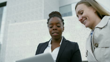 and female colleagues sitting together outdoors, looking at laptop screen and discussing details of business project