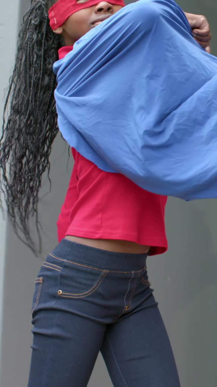 Vertical video: Helper tying strap leading girl posing, twirling cape at wall with red mask to play