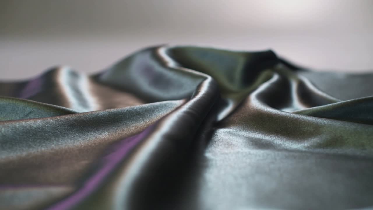 Câmera recuando, tecido de cetim iridescente se movendo sobre a mesa, revelando uma luz de fundo suave.