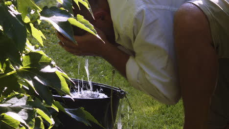 an elderly man, kneeling by a bucket full of rainwater, washes his face on a hot day