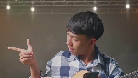 primer plano de un adolescente asiático con una guitarra usando un teléfono inteligente y apuntando hacia un lado mientras está de pie en el escenario