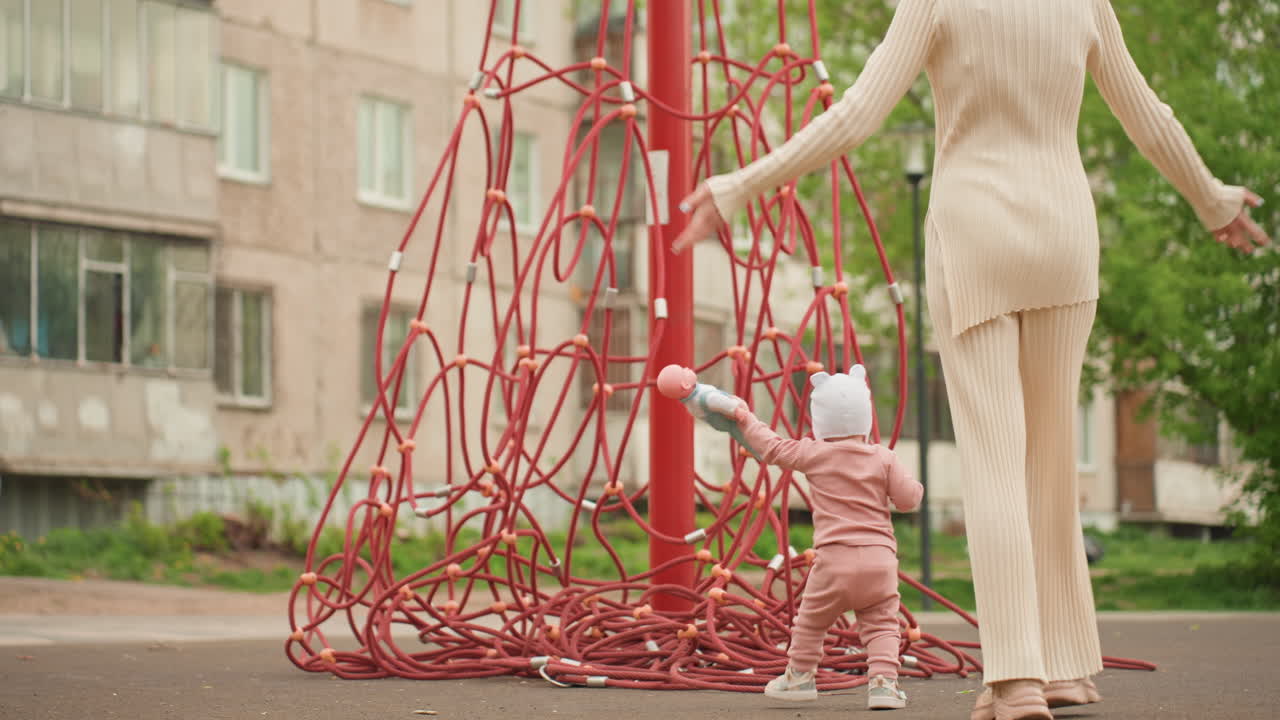 Toddler Walking With Mother Near Rope, Soft Beige Outfit, Guiding Hand Support, Urban Apartment Backdrop, Older Child Passing By, Gentle Encouragement, Learning Balance On Pavement, Family Bonding