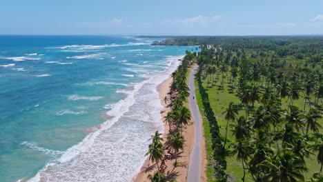 Drone-flight-over-tropical-coastline-with-palm-trees,-sandy-beach-and-waves-on-turquoise-colored-Caribbean-Sea-in-summer---ARROYO-SALADO,-Dominican-Republic