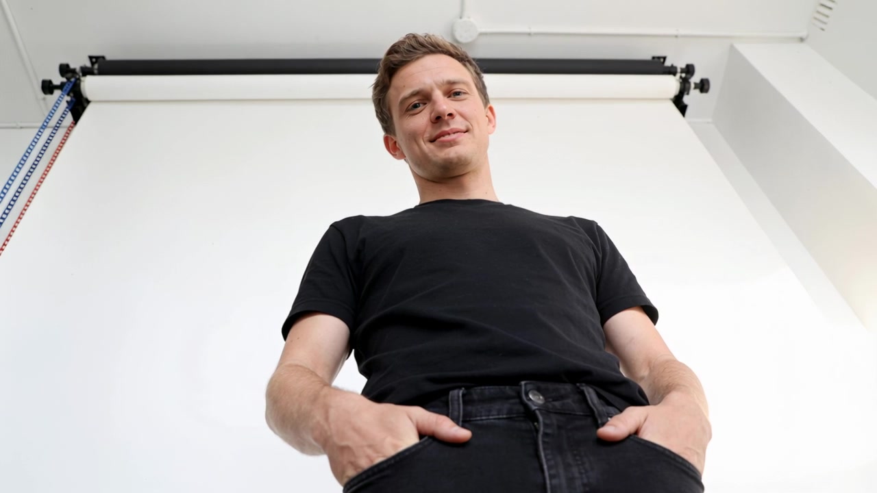 Smiling man easing into neutral expression after first grin in studio with black tee, backdrop roll