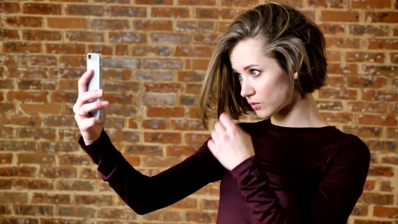 joven hermosa está haciendo selfie en su teléfono inteligente, tocando el cabello, concepto de comunicación, fondo de ladrillo